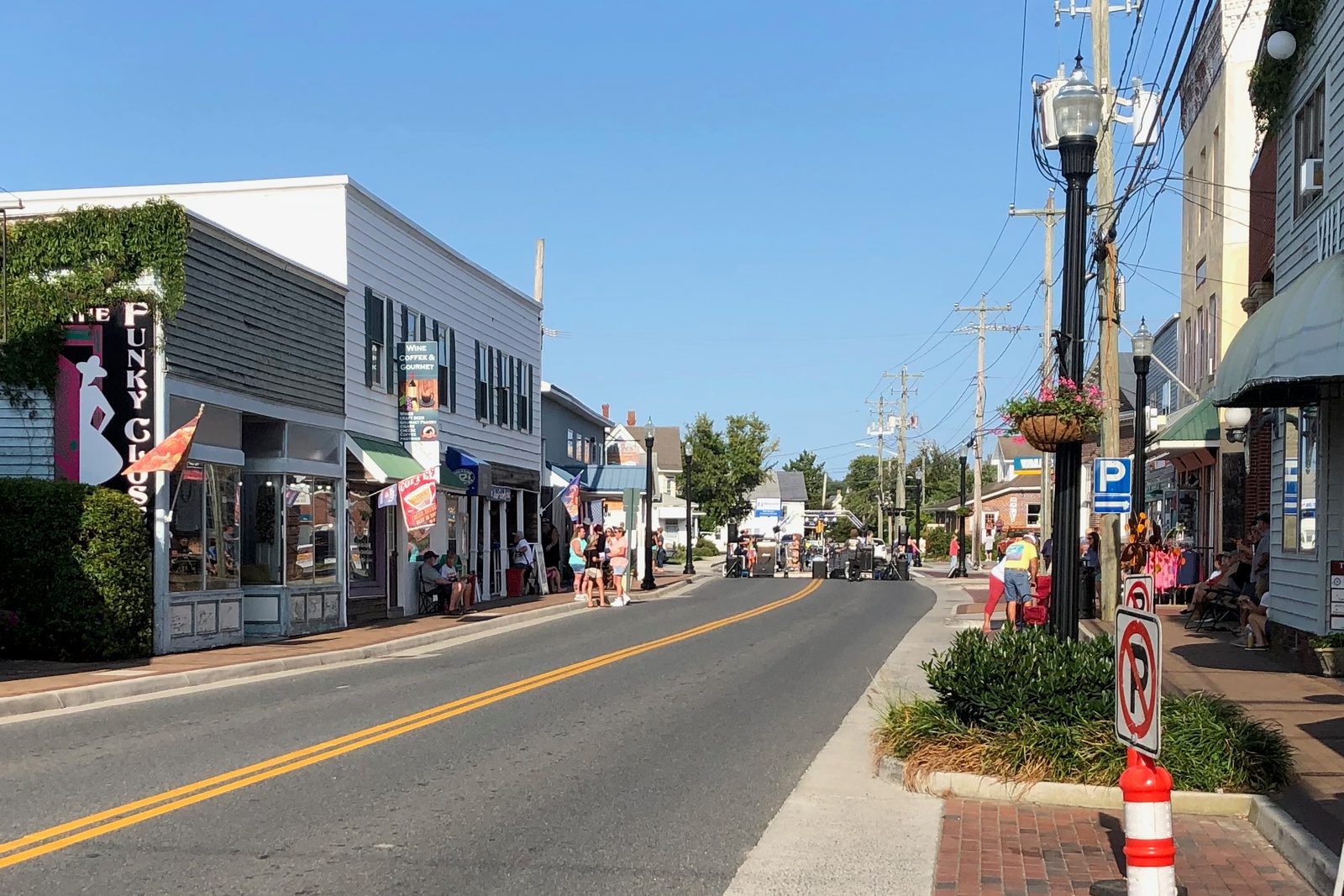 Sunny small-town main street with storefronts, awnings and hanging flower baskets along a two-lane road, with a few people on the sidewalk in the distance